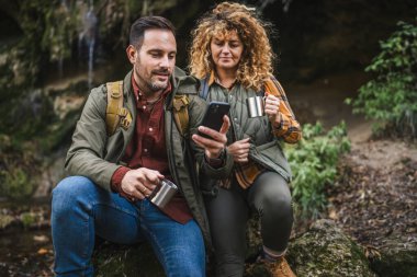 Karı koca yürüyüşçüleri mağaranın önünde oturup metal bardak içip cep telefonu kullanırlar.