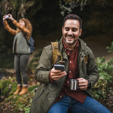Koca yürüyüşçüsü mağaranın önünde oturur fincandan çay içer ve arka planda cep telefonu beyaz karısını kullanır.