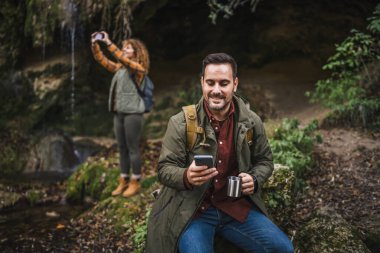 Koca yürüyüşçüsü mağaranın önünde oturur fincandan çay içer ve arka planda cep telefonu beyaz karısını kullanır.
