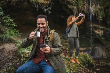 Koca yürüyüşçüsü mağaranın önünde oturur fincandan çay içer ve arka planda cep telefonu beyaz karısını kullanır.