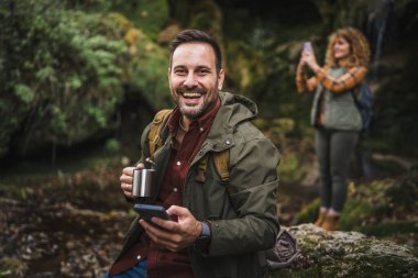 Koca yürüyüşçüsü mağaranın önünde oturur fincandan çay içer ve arka planda cep telefonu beyaz karısını kullanır.
