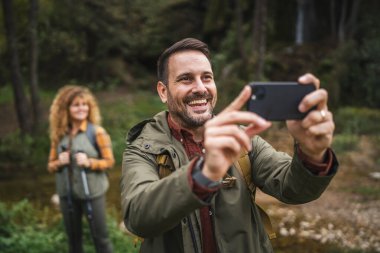 Yetişkin adam yürüyüşçüsü karısı arkasında dururken kendi fotoğrafını çeker.