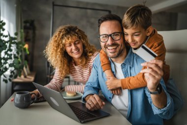 Anne, baba ve oğul laptopta kredi kartıyla alışveriş yaparken, oğul babaları için beklemede.