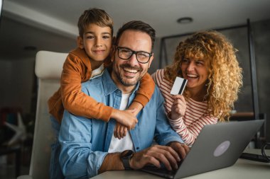 Anne, baba ve oğul laptopta kredi kartıyla alışveriş yaparken, oğul babaları için beklemede.