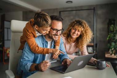 Anne, baba ve oğul laptopta kredi kartıyla alışveriş yaparken, oğul babaları için beklemede.