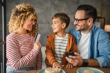Kahvaltıda üç kişilik bir aile cep telefonundan kredi kartıyla online alışveriş yapıyor.