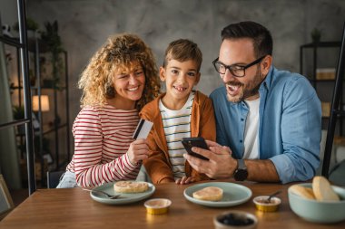 Kahvaltıda üç kişilik bir aile cep telefonundan kredi kartıyla online alışveriş yapıyor.
