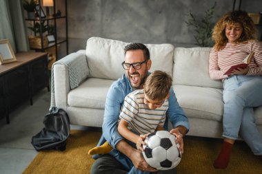 Anne arkalarında kitap okurken baba ve oğul halıya oturup futbol topuyla oynuyorlar.