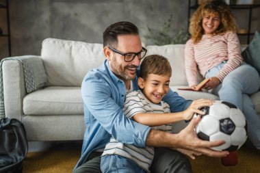 Anne arkalarında kitap okurken baba ve oğul halıya oturup futbol topuyla oynuyorlar.