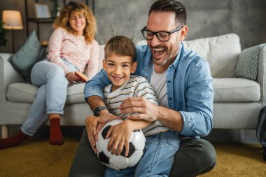 Anne arkalarında kitap okurken baba ve oğul halıya oturup futbol topuyla oynuyorlar.