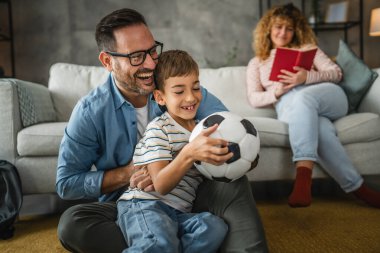 Anne arkalarında kitap okurken baba ve oğul halıya oturup futbol topuyla oynuyorlar.