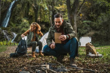 Bir eş, doğayla, çadırı, şelalesi ve sonbahar yapraklarıyla çevrili, mükemmel bir açık hava yaratan, kağıtlarla ateş yakan bir kocanın arkasında eğlenir.