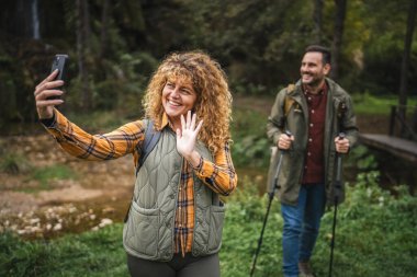 Yetişkin bir kadın yürüyüşçü kocası arkada dururken kendi portresini ya da görüntüsünü çeker.
