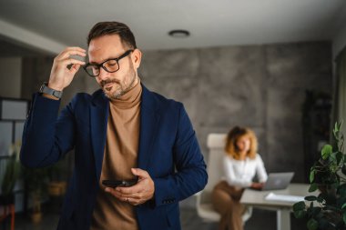 Yorgun yetişkin bir iş adamının portresi arka planda bayan meslektaşıyla ofiste cep telefonu kullanıyor.