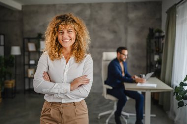 Yetişkin kadın iş kadını portresi arka planda kadın meslektaşı ile ofiste kolları çapraz duruyor.