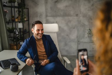 female colleague take a photo with mobile phone of her male colleague in the office clipart