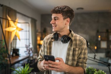 Genç blog yazarının portresi, podcast ekipmanlarının arkasındaki stüdyoda duruyor ve cep telefonu kullanıyor.