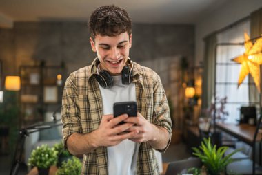 Genç blog yazarının portresi, podcast ekipmanlarının arkasındaki stüdyoda duruyor ve cep telefonu kullanıyor.