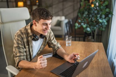 Genç beyaz adam bilgisayarında kredi kartıyla online alışveriş yapıyor.