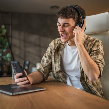 Genç gülen adam evde kulaklıkla ve cep telefonuyla müzik dinliyor.