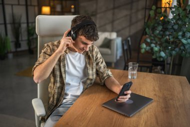 Genç gülen adam evde kulaklıkla ve cep telefonuyla müzik dinliyor.