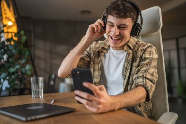 Genç gülen adam evde kulaklıkla ve cep telefonuyla müzik dinliyor.