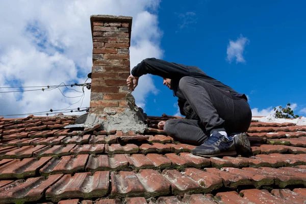 A skilled craftsman on the roof of an old house, using a trowel to repair the chimney. The man sits focused on the task at hand. Sky in background