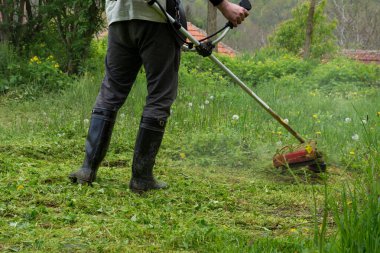 Bir adam bir çim biçme makinesi tutuyor ve bir bahar sabahı bahçesinde uzun otlar kesiyor. Çayırda çalışırken çizmeli çiftçiyi kapat. Tarım kavramı