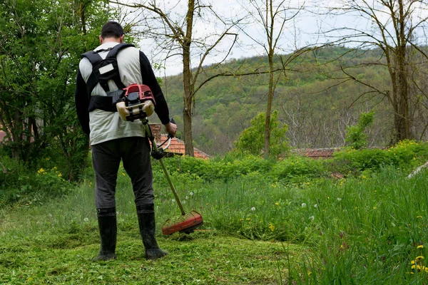 春天的一天 一个男人正在他的农村后院用一只手牵着修剪绿草 这个人穿着靴子 全神贯注地工作 面对着镜头 — 图库照片