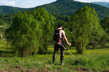 Kırsal kesimde güneşli bir yaz gününde çim biçmek için el makası kullanan bir adam. Sıcaktan gömleksiz