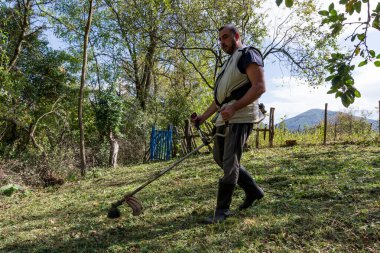 Güneşli bir sonbahar gününde kırsal alanda çim biçme makinesi kullanan bir adam. Arkaplanda, emlak bakımı kavramının vurgulandığı güzel doğal manzara yer alıyor.