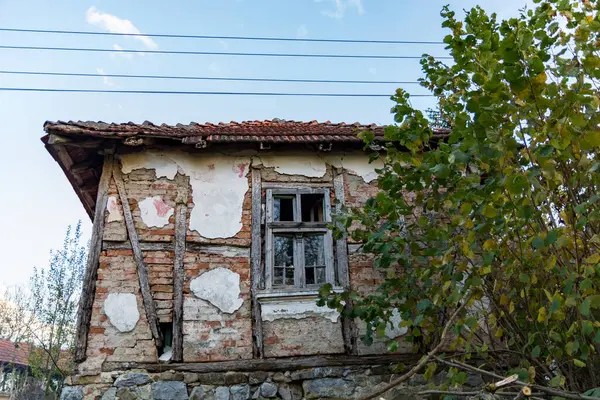 Eski, nostaljik bir köy evi, yıpranmış cephesi kırmızı tuğlaları ve tahta kirişleri ortaya çıkarıyor. Kırık pencere güneşli bir sonbahar gününün resmini çizer.