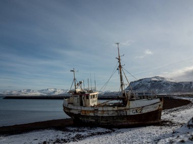 İzlanda 'da karlı bir sahilde terk edilmiş bir balıkçı teknesi, karlı dağlar ve arka planda sakin bir deniz. Hatırlatıcı kış kıyı sahnesi