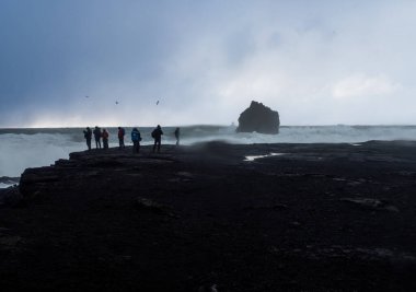Bir grup fotoğrafçı İzlanda 'nın engebeli kıyı şeridinin kenarında duruyor. Fırtınalı gökyüzünün altında tek bir kaya oluşumuna çarpan dramatik dalgaları yakalıyorlar.