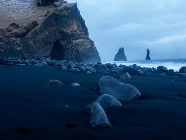 Reynisfjara Sahili 'nin kara kumlarına saçılmış pürüzsüz volkanik taşların yakın çekimi, arka planda Atlantik Okyanusu' ndan yükselen bazalt kayalıklar ve Reynisdrangar deniz yığınları.