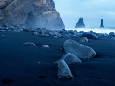 Reynisfjara Sahili 'nin kara kumlarına saçılmış pürüzsüz volkanik taşların yakın çekimi, arka planda Atlantik Okyanusu' ndan yükselen bazalt kayalıklar ve Reynisdrangar deniz yığınları.
