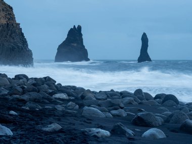 Reynisfjara Sahili alacakaranlıkta simgesel Reynisdrangar deniz yığınları okyanustan yükseliyor, karanlık bazalt sütunlar tarafından çerçeveleniyor ve kasvetli bir gökyüzünün altında volkanik kayalar saçılıyor.