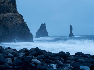 Reynisfjara Sahili, alacakaranlıkta simgesel Reynisdrangar deniz yığınlarıyla çalkantılı dalgalara karşı duruyor, bazalt sütunlar ve dağınık volkanik kayalar tarafından çerçevelenmiş.