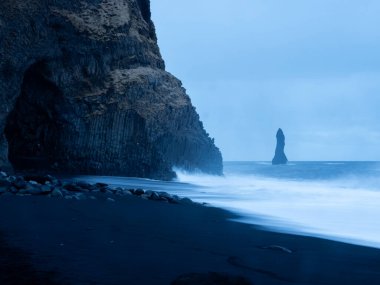 Reynisfjara Sahili alacakaranlıkta, ikonik Reynisdrangar deniz yığını ve bazalt sütun kayalıkları, köpüklü dalgaların siyah volkanik kuma çarpmasıyla