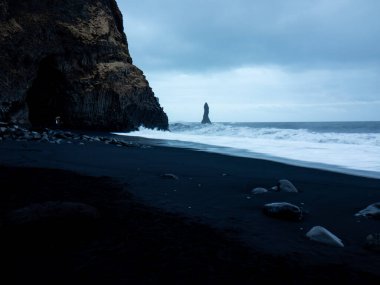 Reynisfjara Sahili, alacakaranlıkta dramatik siyah kum, bazalt mağarası, engebeli uçurumlar ve çalkantılı Atlantik Okyanusu 'ndan yükselen ikonik Reynisdrangar deniz yığınlarıyla dolu.