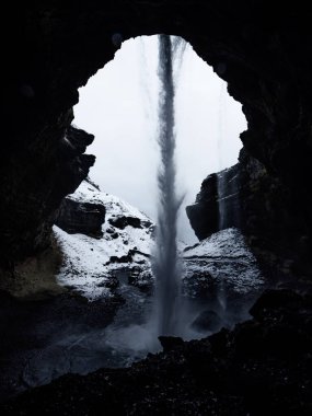 Arkadan görülen Kvernufoss şelalesi, karanlık bir mağara açıklığıyla çerçevelenmiş. Şelale gibi akan su, kar ve dışarıdaki ışıkla tamamen zıtlaşıyor ve dramatik, mistik bir sahne yaratıyor.