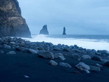 Reynisfjara Sahili alacakaranlıkta bazalt sütunları ve Reynisdrangar deniz yığınlarıyla. Siyah kumsal, dramatik mavi alacakaranlık gökyüzünün altında pürüzsüz volkanik kayalarla dağılmış.