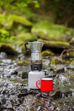 Coffee pot and metal cup on a large stone against the background of a mountain river. Coffee pot on a stone slab against a blurred background of nature. clipart