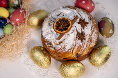 Easter Kulich decorated with cinnamon and dried orange, with golden Easter eggs next to it on a light background