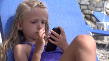 Kid Playing Smartphone on Sunbed on Beach, Child Browsing Internet on Smart Phone on Seashore, Girl with Touchscreen Telephone