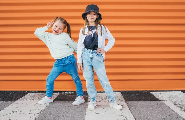 两个小女孩在靠近橙色墙的城市人行横道上 在镜头前摆姿势 给孩子们穿上时髦的衣服 快乐的现代童年概念形象 — 图库照片