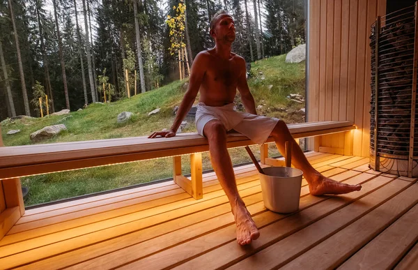 Relaxing man wrapped white towel sitting on the wooden bench in Hot Finnish sauna with a huge wide window with green forest view and enjoying pleasant healthy body care temperature treatment.