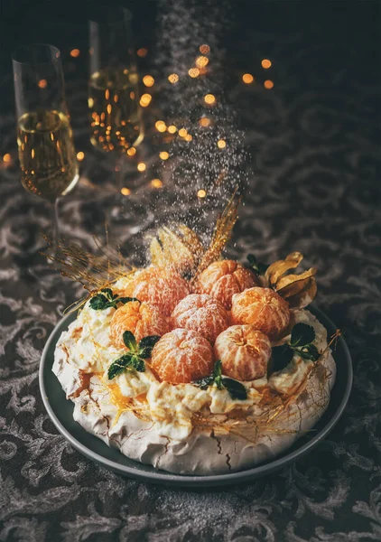 Pavlova cake low light still life pouring with powder sugar meringue-based dessert named after the Russian ballerina decorated with fresh mandarins, caramel, mint, physalis with Christmas lights
