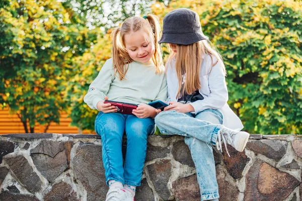 两个笑着的妹妹女孩坐在秋天的公园里浏览着她们的智能手机装置 粗心大意的童年时光与现代技术观念形象 — 图库照片
