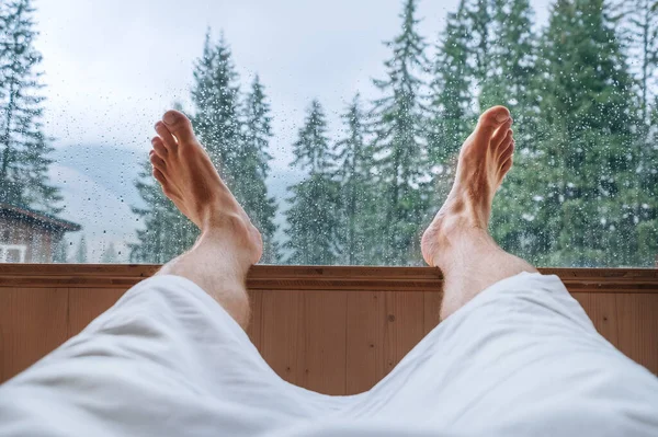 Man's legs POV relaxing man wrapped towel lying in Hot Finnish sauna with a huge wide window with green spruce forest view and enjoying pleasant healthy body care temperature treatment.
